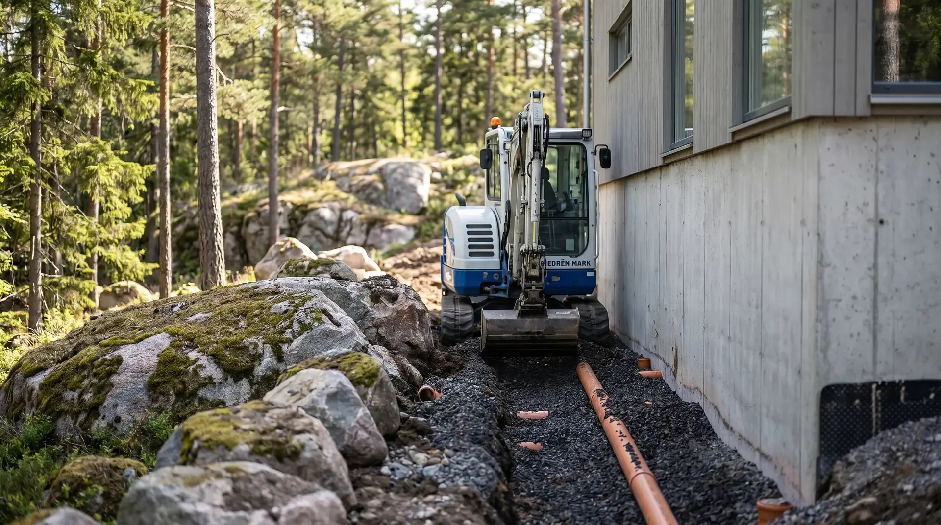 En minigrävare arbetar intill en husvägg omgiven av skog och stenar.